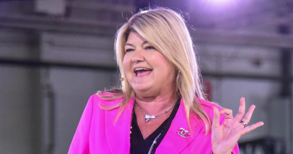 Pessoa em pé atrás de um púlpito transparente, vestindo um blazer rosa-choque sobre uma blusa preta. O rosto da pessoa está desfocado. Ela faz um gesto de 'OK' com a mão direita. (IA)